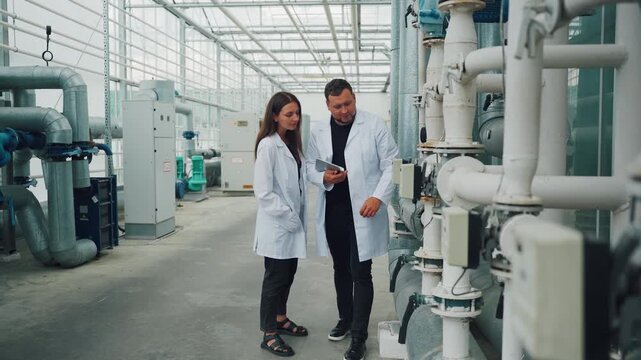 Inspecting greenhouse equipment. Engineers analyze heating system components. Two professionals examine greenhouse heating infrastructure while discussing repair strategies and modern technology