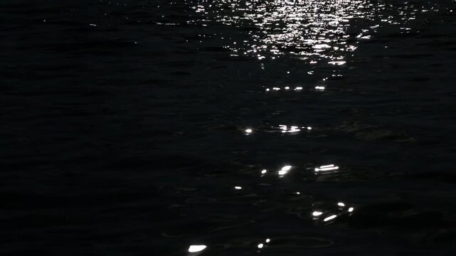 The dark surface of ocean water at night is shot from above as bright light moves from the center to the right side, creating a dynamic pattern of light reflecting on the black water.