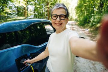 Naklejka premium Senior woman with short gray hair and glasses smiles while charging an electric vehicle on a forest road, surrounded by lush green trees and sunlight filtering through