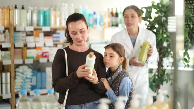 Interested woman in thirties reading label on shampoo, checking for baby-safe ingredients while choosing hair care product for tween daughter. Friendly pharmacist offering options in background. High