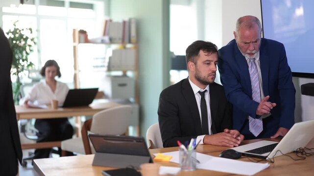 Strict mature man boss scolding new adult man office worker for improper operation on laptop in modern office