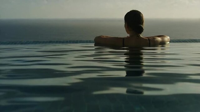 Gazing woman wearing dark straps relaxing on infinity pool edge sea, sun and breeze moving ripples