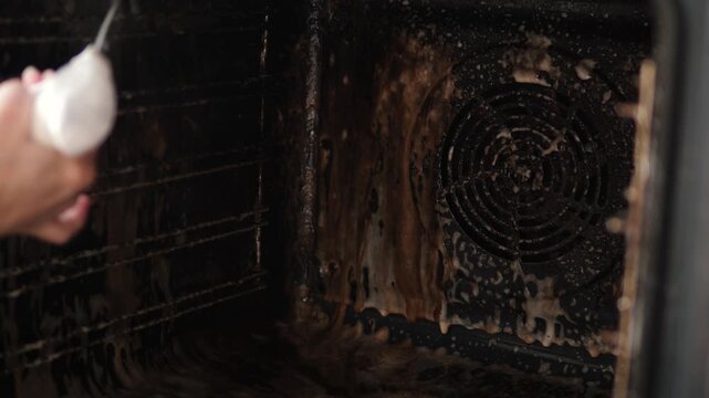 Close-up of spraying a cleaning product onto the heavily soiled back wall of an oven. This is the process of removing stubborn grease and burnt-on grime during a deep kitchen clean