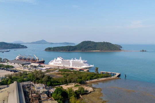 Docked at a vibrant port, a majestic cruise ship and a robust cargo vessel sit in clear blue water on a sunny day. Nearby buildings and vehicles are visible