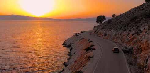 Naklejka premium DRONE: Flying behind a big gray car driving along the coastal road at sunrise.