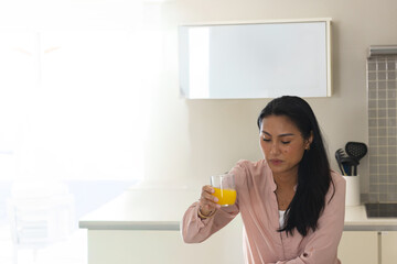 Middle-aged Asian woman sitting at kitchen counter holding clear glass of orange juice, copy space © wavebreak3