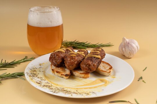 Grilled Romanian mici skinless sausages drizzled with mustard, served on toasted bread slices next to a glass of cold beer and a garlic bulb.