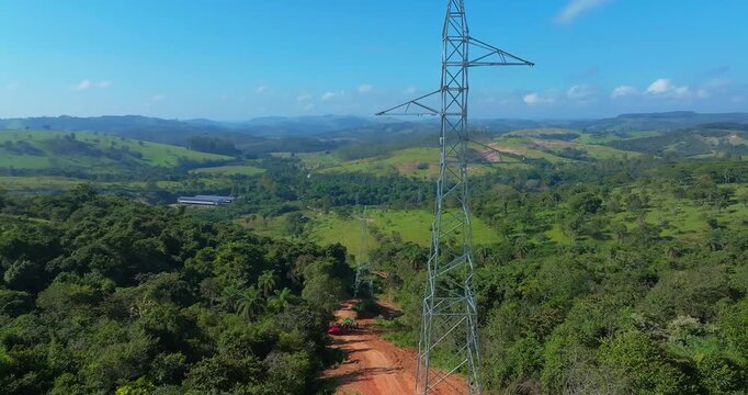 Electrical transmission tower crossing tropical rural landscape near Santo Ant&ocirc;nio do Monte, Minas Gerais, Brazil with rolling hills, forest fragments, pastureland and red dirt road, slow forward fly