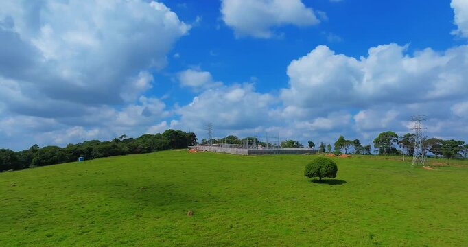 High voltage electrical substation surrounded by green cattle pasture and tropical rural hills near Santo Ant&ocirc;nio do Monte, Minas Gerais, Brazil beneath large cumulus cloud formations, drone forward