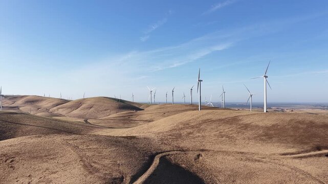 Tracking drone view of a wind farm on rolling brown hills, showing wind turbines spinning and generating clean renewable energy against a clear blue sky on a sunny day at Altamont Pass in California