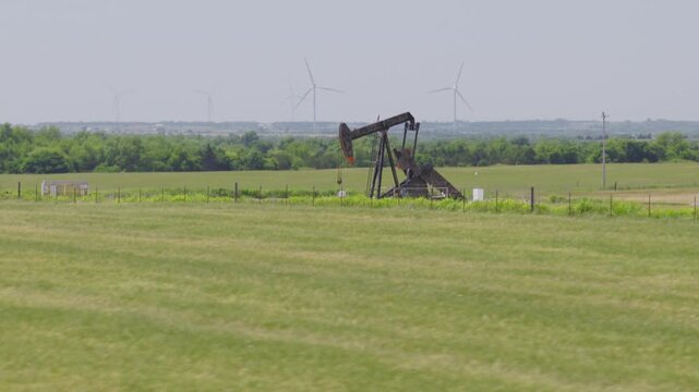 Drone 4K of a green field with an oil pump jack and wind turbines in the background.