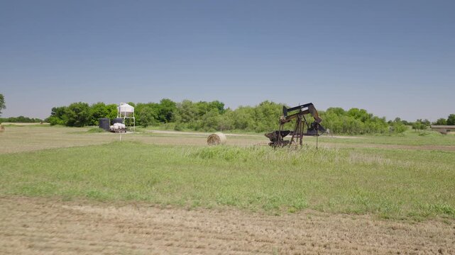 Drone 4K of a field of hay bails surrounding an oil pump jack in the spring and summer.