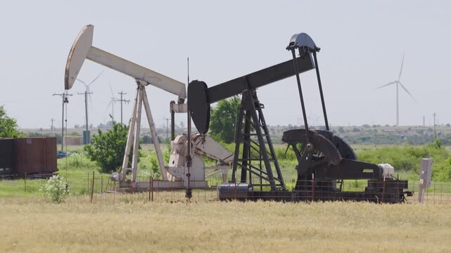 Drone 4K of a wheat field with two oil pump jacks and wind turbines in the background.
