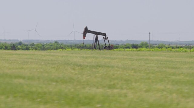 Drone 4K of a green field with an oil pumpjack and wind turbines in the background.