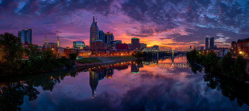 A vibrant sunset casts colorful reflections on the calm water, mirroring the silhouetted cityscape and its lit structures. The sky is ablaze