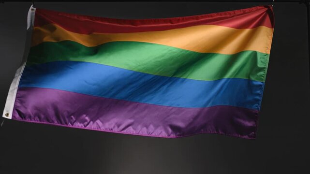 Colorful pride flag with horizontal stripes of red, orange, yellow, green, blue, and purple waving gently against a dark background, showcasing vibrant hues and fabric texture