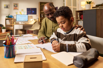 Naklejka premium Black father helping school age son writing homework at table, supporting home learning and parental involvement. Ideal for education, tutoring, parenting, child development campaigns