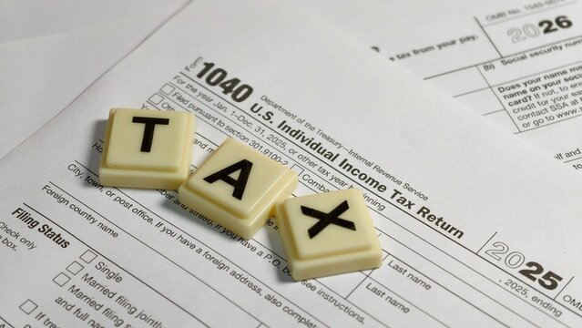 An IRS Form 1040 U.S. Individual Income Tax Return for 2025 lies beneath another tax document marked 2026, with three alphabet letter tiles spelling out "TAX" placed diagonally on top.
