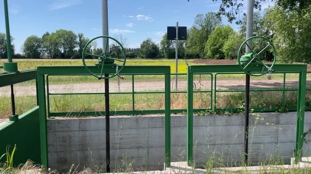 Water Gate Control: Industrial water gates with large green wheels stand ready to manage water flow in a rural setting, with fields and trees visible under a clear blue sky.