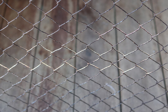 abstract view of a rusty steel wire mesh fence against a blurred wooden background. This image targets commercial security themes, prison boundaries, industrial safety partitions, construction site