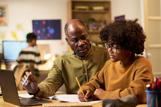 Black senior man guiding Black teenager studying with laptop and notebook at home. Family learning support and homeschooling concept for education campaigns and parenting content