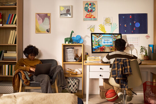 African American teenager studying on laptop while younger boy attending online math class at desk in shared room, showing home education and sibling learning for edtech campaigns