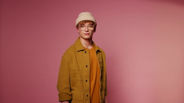 Portrait of a young man in a beanie, glasses, and an overshirt standing calmly against a pink background, looking at the camera.