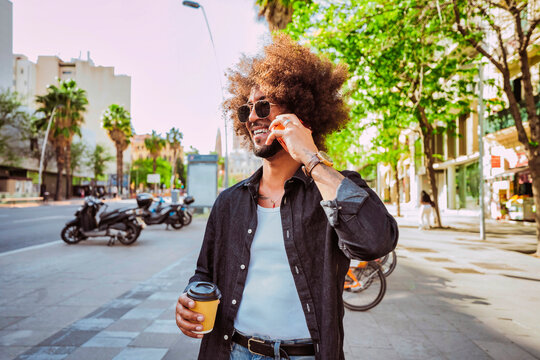 Latin american man with afro hair smiling while enjoying his coffee and talking on the phone in an urban setting.