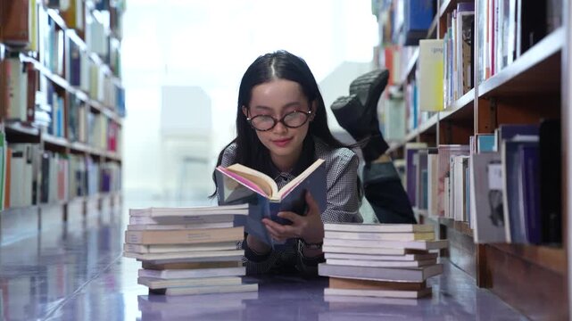 Young Asian woman and teenage student with glasses lying on the floor reading a book in the library. Female college student studying with a stack of textbooks near bookshelves.