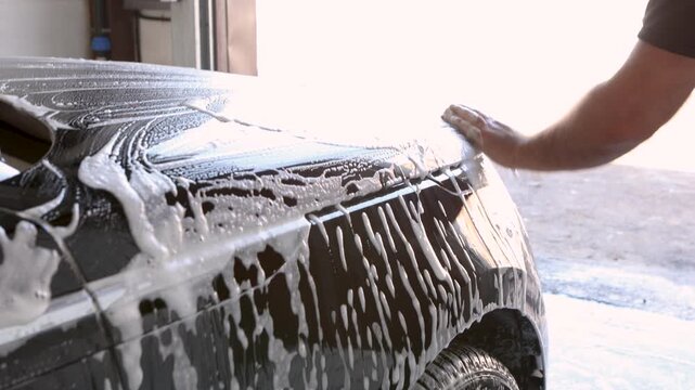 Car wash, close up of male hand wiping black car with sponge full of foam at indoor station. Soap texture and smooth strokes remove dirt, professional auto detailing and cleaning service