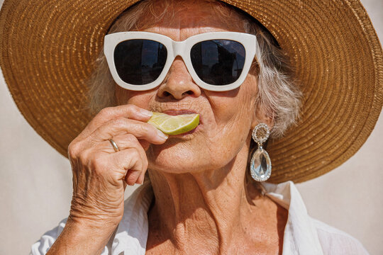 Stylish elderly woman in a wide brim straw hat and white sunglasses tasting a sour lime slice against a neutral background during a bright summer vacation.