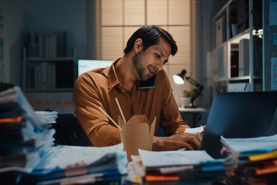 Young Latin businessman taking urgent phone call while reading accounting document on laptop at desk in dark night office. Multitasking overtime, overworked deadline, tired paperwork rush concept.