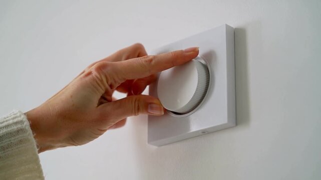 Reaching right hand manicured nails knit-sleeve turning wall dial at home, adjusting thermostat