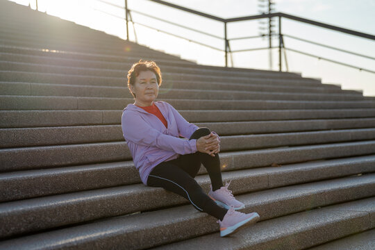Senior Asian woman resting on outdoor steps after morning run, active lifestyle concept