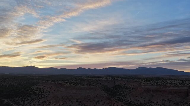 4K drone footage features a powerful atmospheric transition at sunset. Layers of glowing colorful clouds move and diffuse over a dark mountain skyline and arid desert mesas.