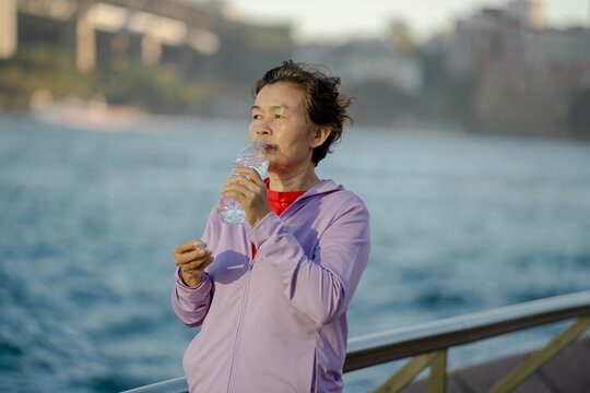 Senior Asian woman drinking water after outdoor exercise by harbour waterfront, healthy lifestyle concept