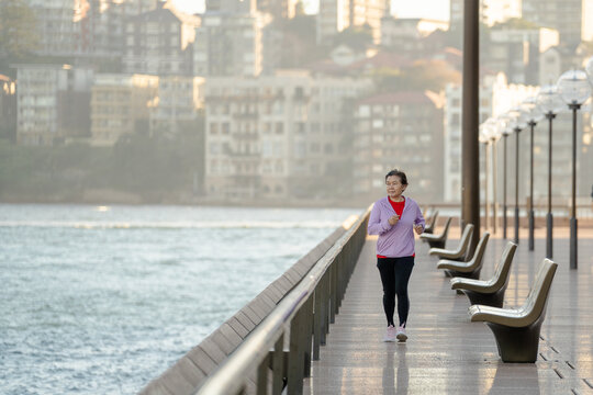 Active senior Asian woman jogging along waterfront promenade at sunrise, healthy lifestyle concept