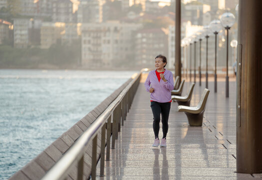 Active senior Asian woman jogging along waterfront 
promenade at sunrise, healthy lifestyle concept