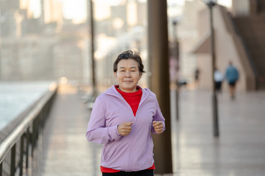 Close up Active senior Asian woman jogging along waterfront promenade at sunrise, healthy lifestyle concept