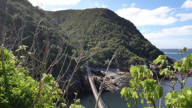 A breathtaking aerial view of a partial suspension bridge, rugged cliffs, the ocean, and lush forests creates a dramatic landscape at Storm&rsquo;s River Mouth in Tsitsikamma National Park, South Africa.