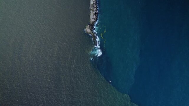 Top-down drone shot showing the color contrast between brown river water and blue ocean. Aerial tracking along a concrete breakwater dividing two distinct water bodies