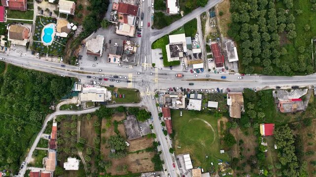 Wide aerial top-down perspective of a busy street intersection and crossroad traffic in the rural settlement of Roda, North Corfu, Greece. Drone overhead view of village houses and local roads.