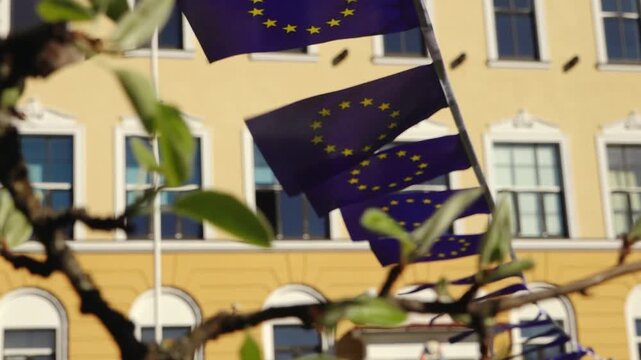 Blurred foreground leaves frame European Union flag ribbons hanging in front of a yellow building, suggesting an outdoor civic or cultural event.
