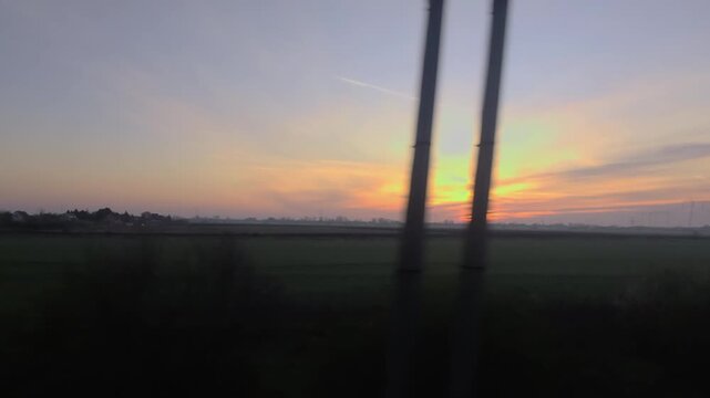 A cinematic static shot captured through a train window, image_2c3da0.png features the golden sun cresting the horizon. The warm morning light spreads across the flat rural landscape and silhouetted t