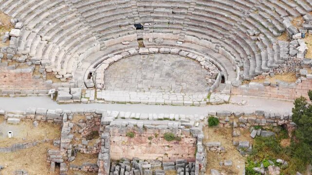 Aerial view zooming out from the ancient theatre of Delphi, a famous archaeological site in Greece, revealing the semicircular auditorium, orchestra, and the surrounding historic ruins