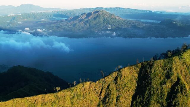 Volcanic caldera lake in Indonesia with crater landscape and steep mountain slopes, sunlit terrain with clouds and Mount Batur in the background, push-in aerial drone footage