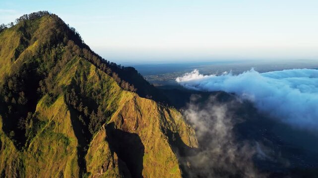 Mount Abang Bali volcanic mountain ridge with steep slopes and rugged green terrain in golden hour light across highland landscape with deep shadows and soft clouds over Lake Batur aerial drone view