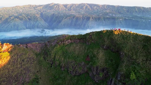 Adventurers watching sunrise from steep volcanic ridgeline above clouds and mist filled valley with rugged mountain peaks in scenic Bali landscape right lateral drone shot