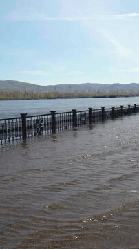 Flooded urban river embankment with partially submerged fence during spring high water and seasonal flooding on Yenisei River