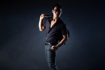 Fototapeta premium An Asian man in all-black formal attire poses against a dark background with dramatic studio lighting, holding his jacket and wearing stylish glasses.